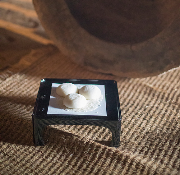 Three freshly pounded round mochi placed on a small stand on a straw mat, with an inverted wooden mortar (usu) in the background on the earthen floor